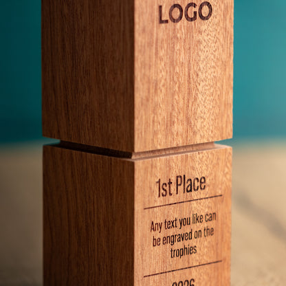 Close-up of a wooden trophy block with engraved text reading 'LOGO' and '1st Place' on a warm wood surface against a teal background.