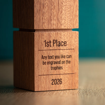 A close-up of a wooden trophy block engraved with '1st Place', placeholder text, and '2026' on a wooden surface against a teal background.