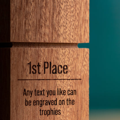 Close-up of a wooden trophy block engraved with the text '1st Place' and 'Any text you like can be engraved on the trophies' against a teal background.