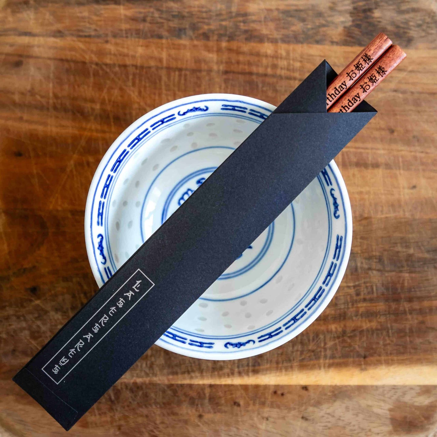 A pair of Japanese-style Chopsticks in a paper pouch, colour rose wood. The chopsticks are laser engraved and personalised with either a name or a phrase in English and are translated into either Chinese or Japanese.