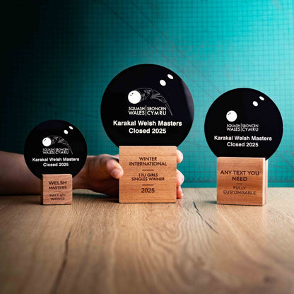 Three squash trophies with custom text and logos, on eco-friendly wooden bases, with a black Perspex squash ball mounted on top, featuring fully customisable areas for text and logos with a hand holding the large trophy to show a perspective of size.