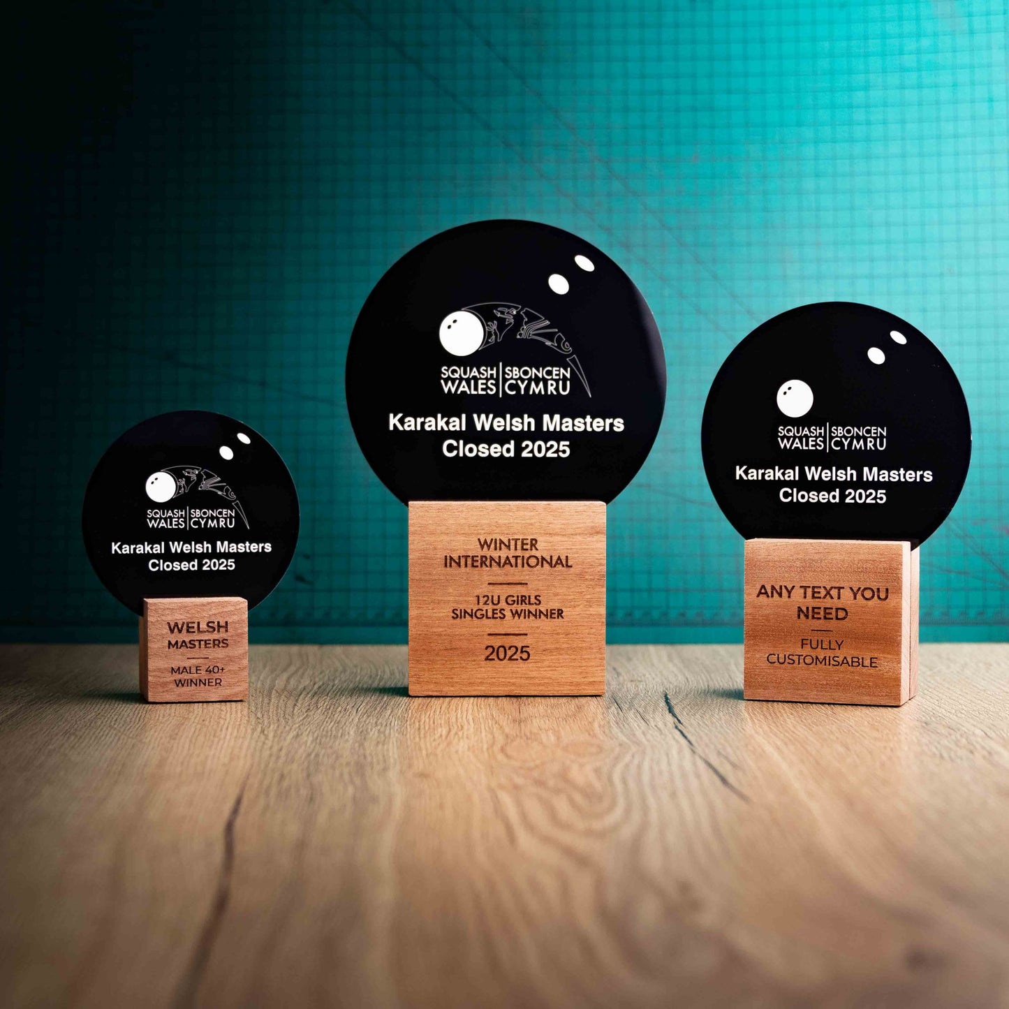 Three squash trophies with custom text and logos, on eco-friendly wooden bases, with a black Perspex squash ball mounted on top, featuring fully customisable areas for text and logos.