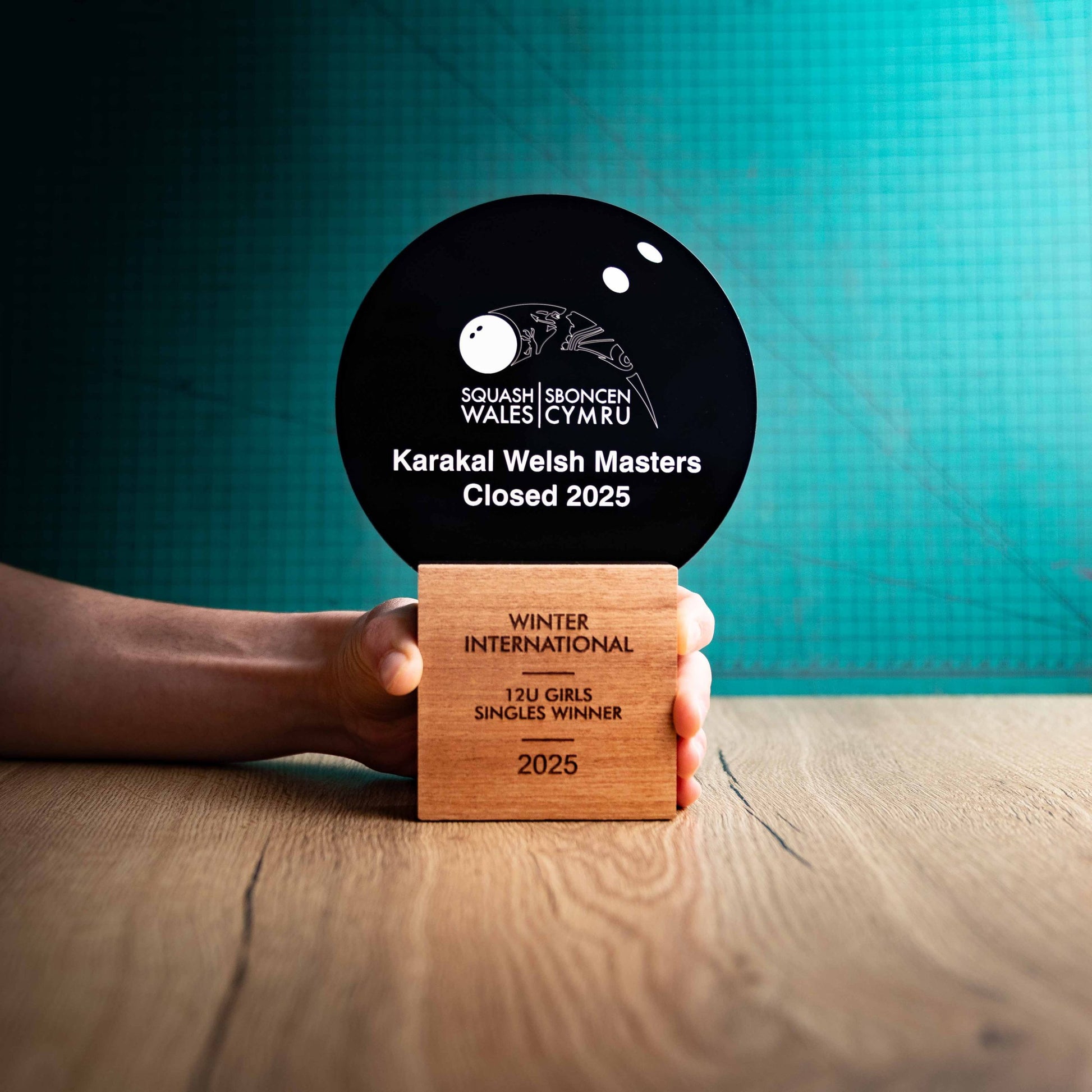 A hand holding a large Squash trophy with custom text and logos, on an eco-friendly wooden base, with a black Perspex Squash ball mounted on top, featuring fully customisable areas for text and logos.
