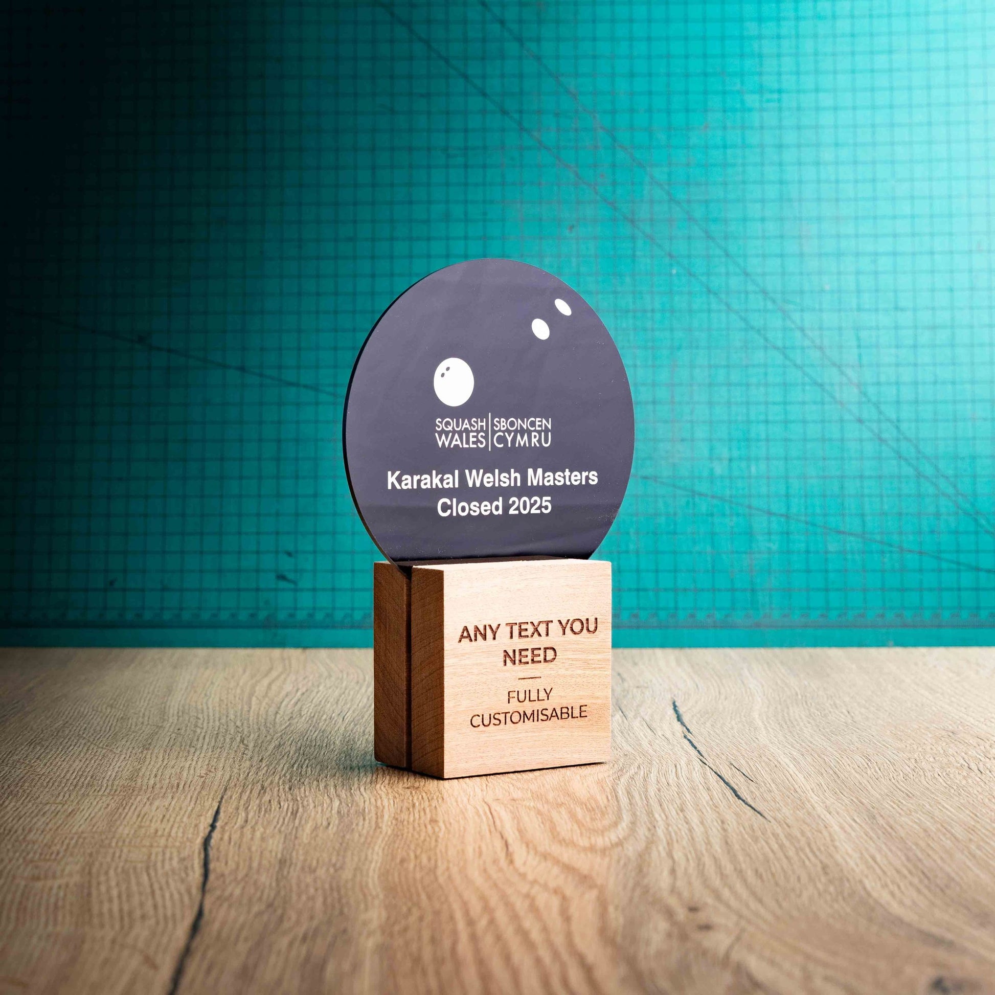 The side view of a medium Squash trophy with custom text and logos, on an eco-friendly wooden base, with a black Perspex Squash ball mounted on top, featuring fully customisable areas for text and logos.