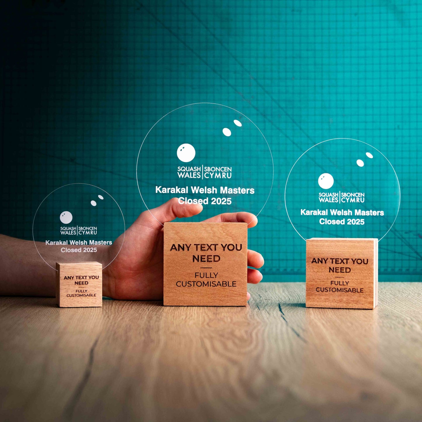 Three squash trophies with custom text and logos, on eco-friendly wooden bases, with a clear Perspex squash ball mounted on top, featuring fully customisable areas for text and logos with a hand holding the large trophy to show a perspective of size.