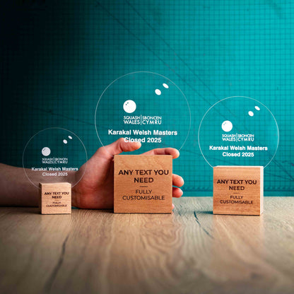 Three squash trophies with custom text and logos, on eco-friendly wooden bases, with a clear Perspex squash ball mounted on top, featuring fully customisable areas for text and logos with a hand holding the large trophy to show a perspective of size.