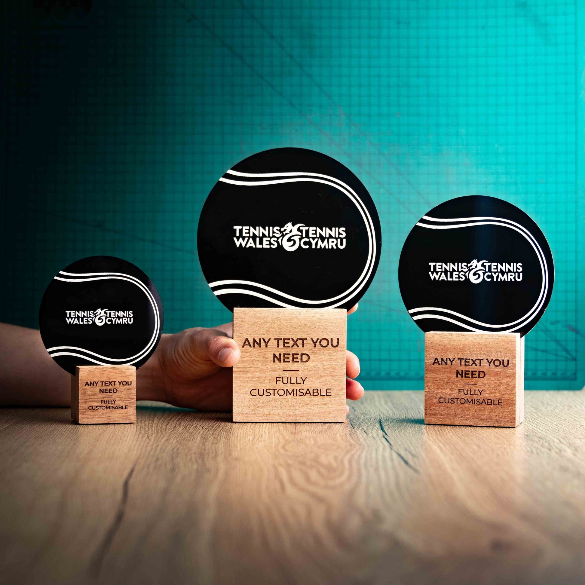 Three Tennis trophies with custom text and logos, on eco-friendly wooden bases, with a black Perspex Tennis ball mounted on top, featuring fully customisable areas for text and logos, with a hand holding the large trophy to give a perspective of the size.