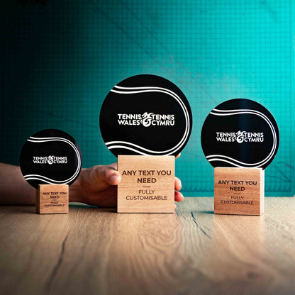 Three Tennis trophies with custom text and logos, on eco-friendly wooden bases, with a black Perspex Tennis ball mounted on top, featuring fully customisable areas for text and logos, with a hand holding the large trophy to give a perspective of the size.
