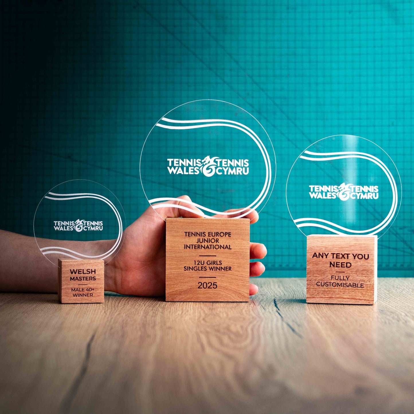 Three Tennis trophies with custom text and logos, on eco-friendly wooden bases, with a clear Perspex Tennis ball mounted on top, featuring fully customisable areas for text and logos, with a hand holding the large trophy to give a perspective of the size.