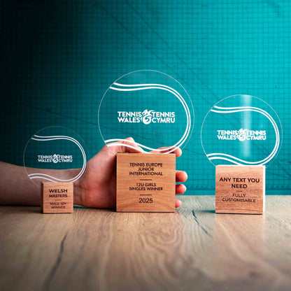 Three Tennis trophies with custom text and logos, on eco-friendly wooden bases, with a clear Perspex Tennis ball mounted on top, featuring fully customisable areas for text and logos, with a hand holding the large trophy to give a perspective of the size.