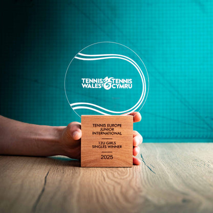 A large Tennis trophy with custom text and logos, on an eco-friendly wooden base, with a clear Perspex Tennis ball mounted on top, featuring fully customisable areas for text and logos, being held to give a size perspective.