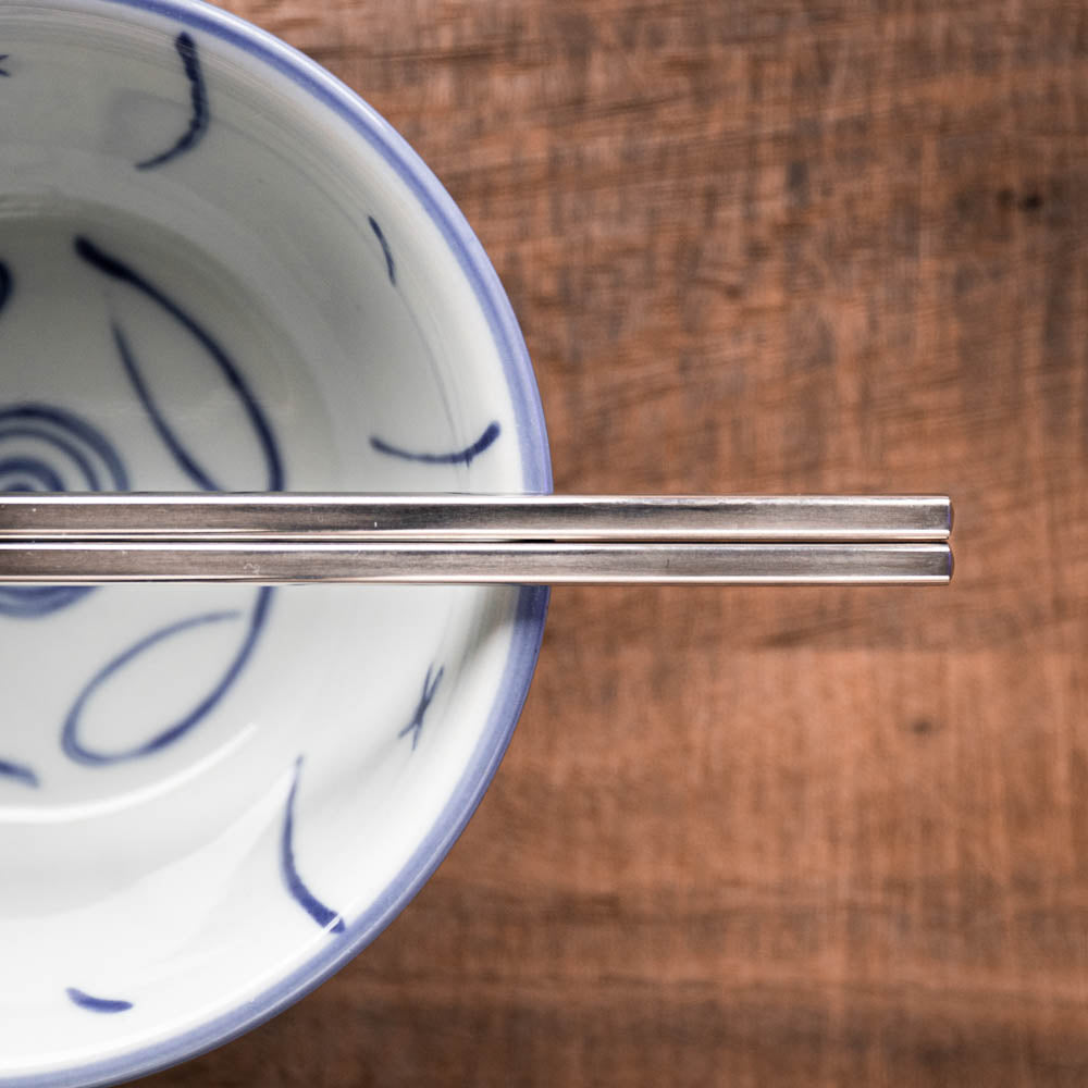 Pair of titanium metal chopsticks on a white ceramic bowl with blue patterns on a wooden surface