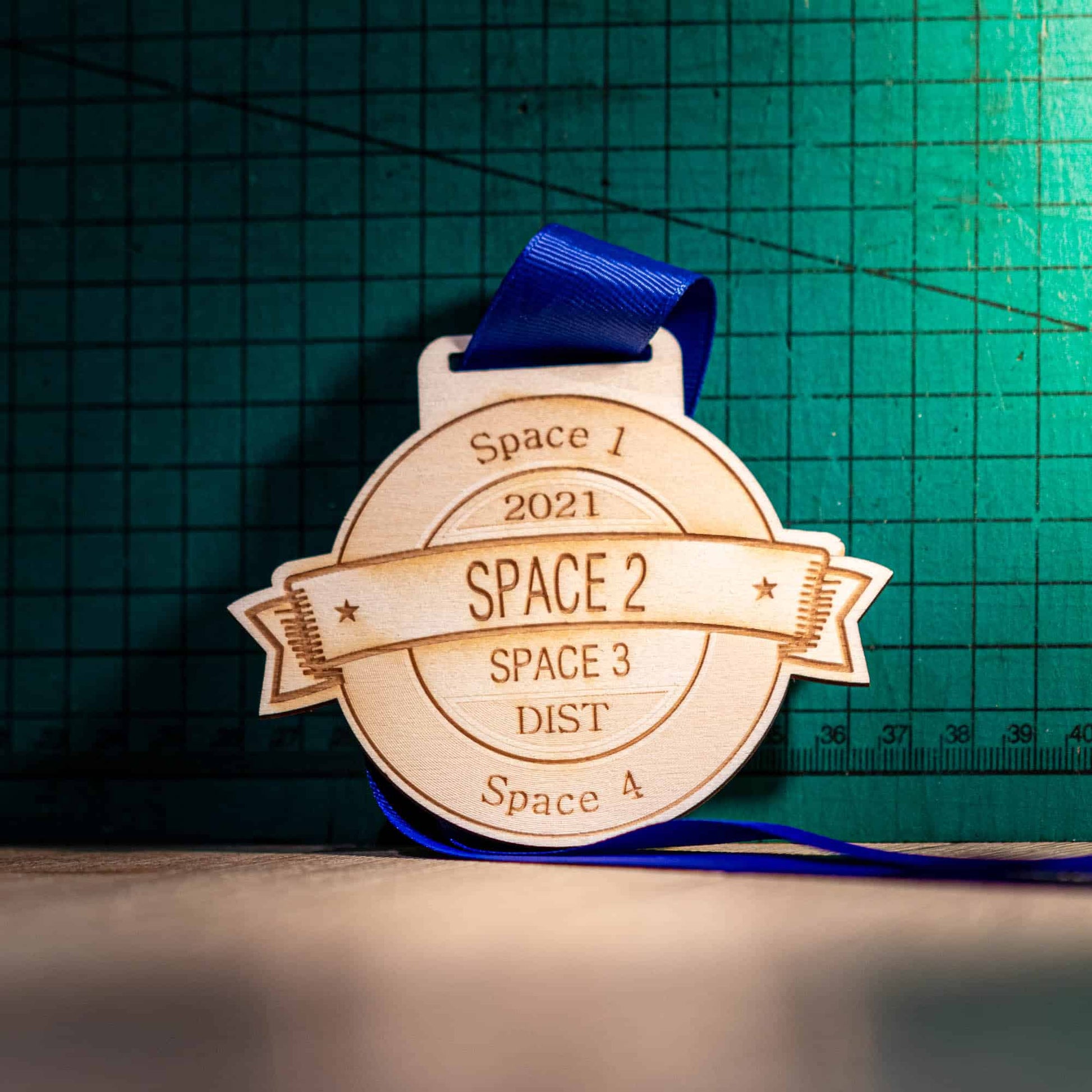 A wooden medal with a blue ribbon, featuring engraved text that includes 'Space 1 2021', 'Space 2', 'Space 3', 'DIST', and 'Space 4'. The medal is circular with a natural wood edge and a branded logo at the top.